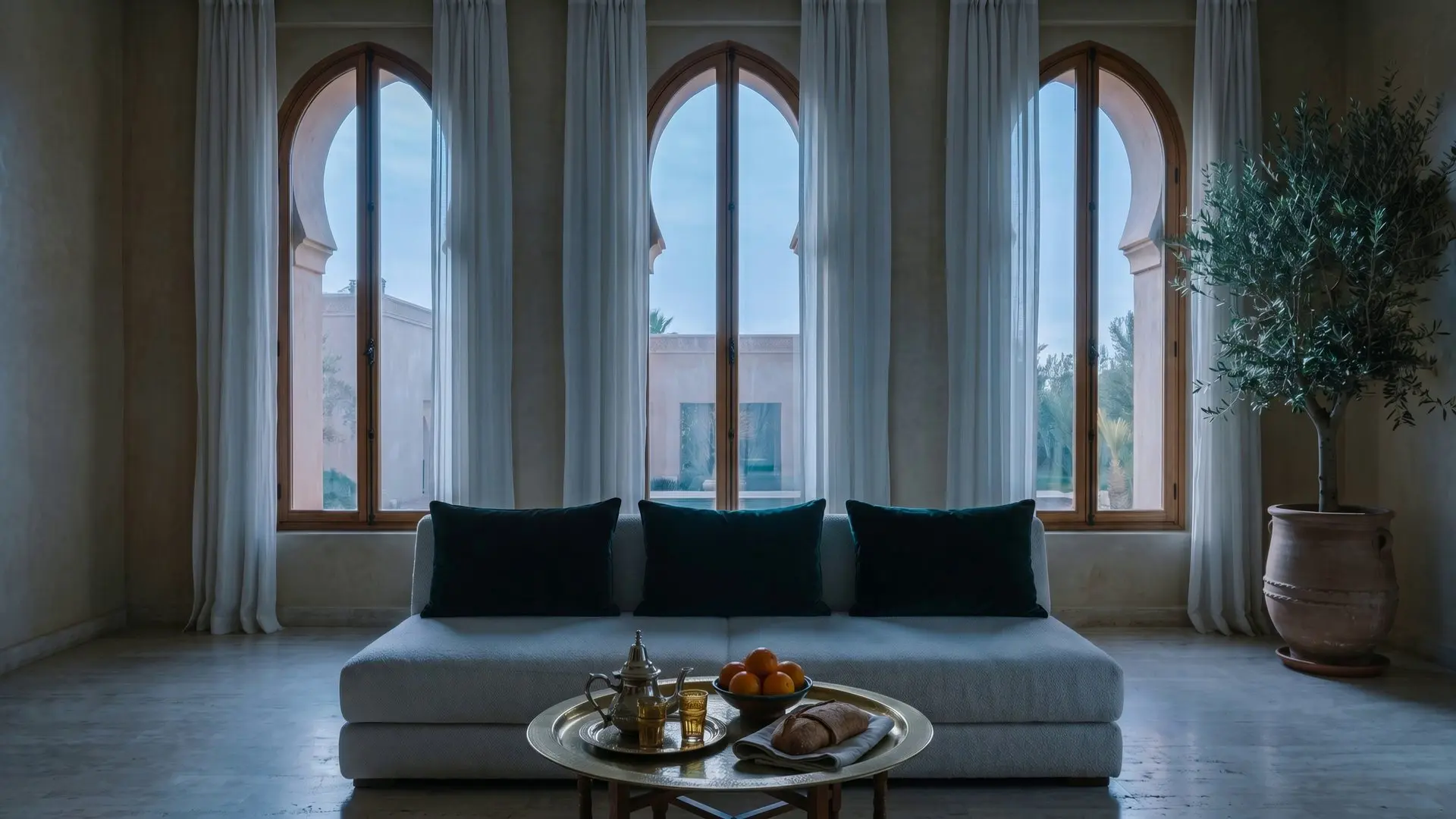 A Moroccan salon at dawn: three arched windows, a cream sofa, a brass tray set for breakfast